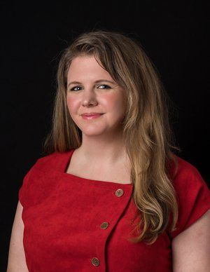 A smiling person with long light brown hair wearing a red shirt against a plain black background.