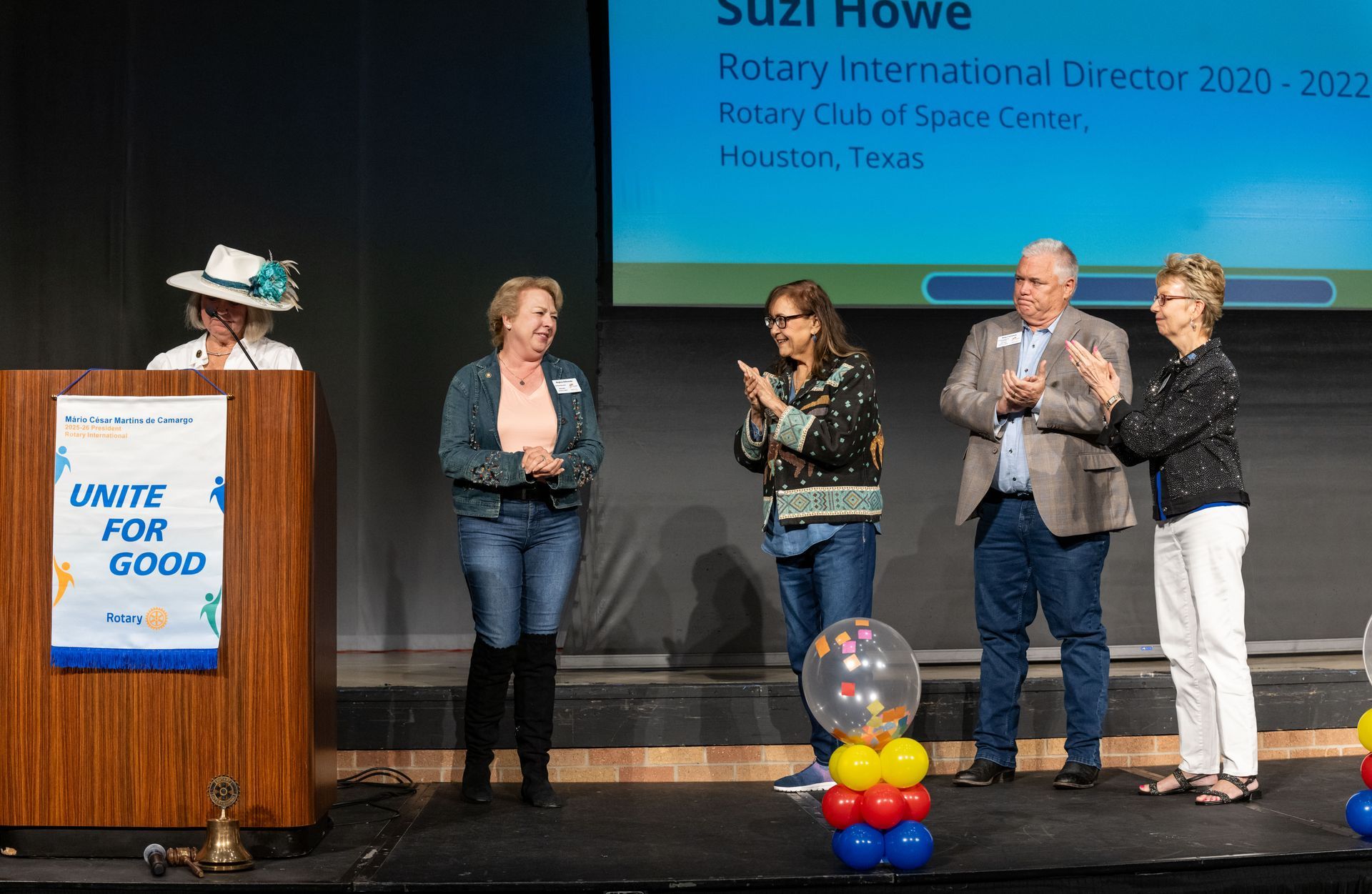 Five people on a stage at a Rotary event, with one speaking at a podium labeled 