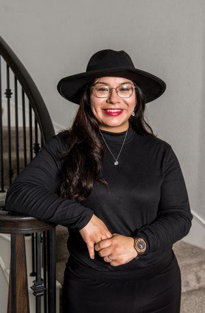 A smiling person in a black hat, mock-neck shirt, and glasses leans against a dark wooden stair railing.