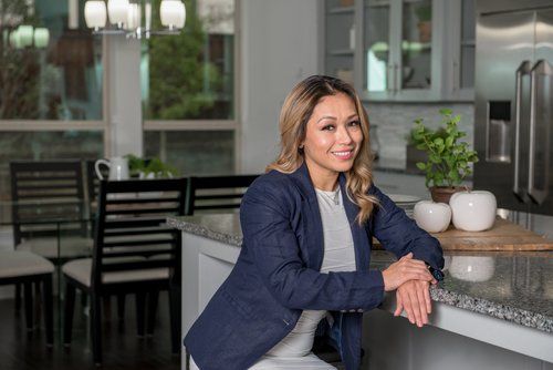 A smiling professional sits at a kitchen island, wearing a dark blazer over a light shirt in a contemporary home setting.