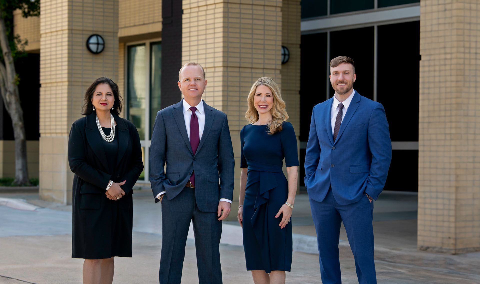 Four professionals in business attire pose for a group photo in front of a brick building.