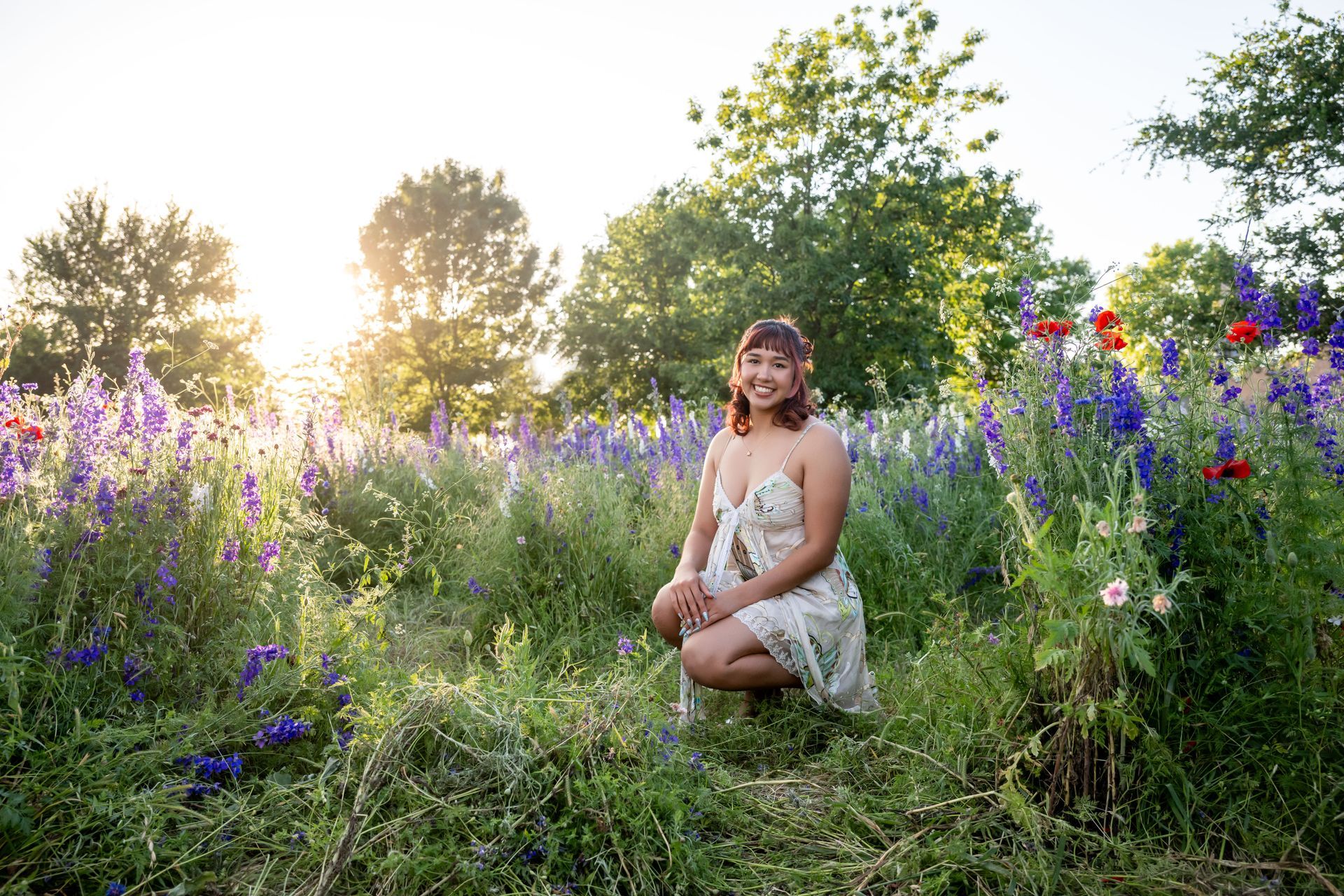 A person in a light sundress crouches in a field of purple flowers and greenery during sunset.