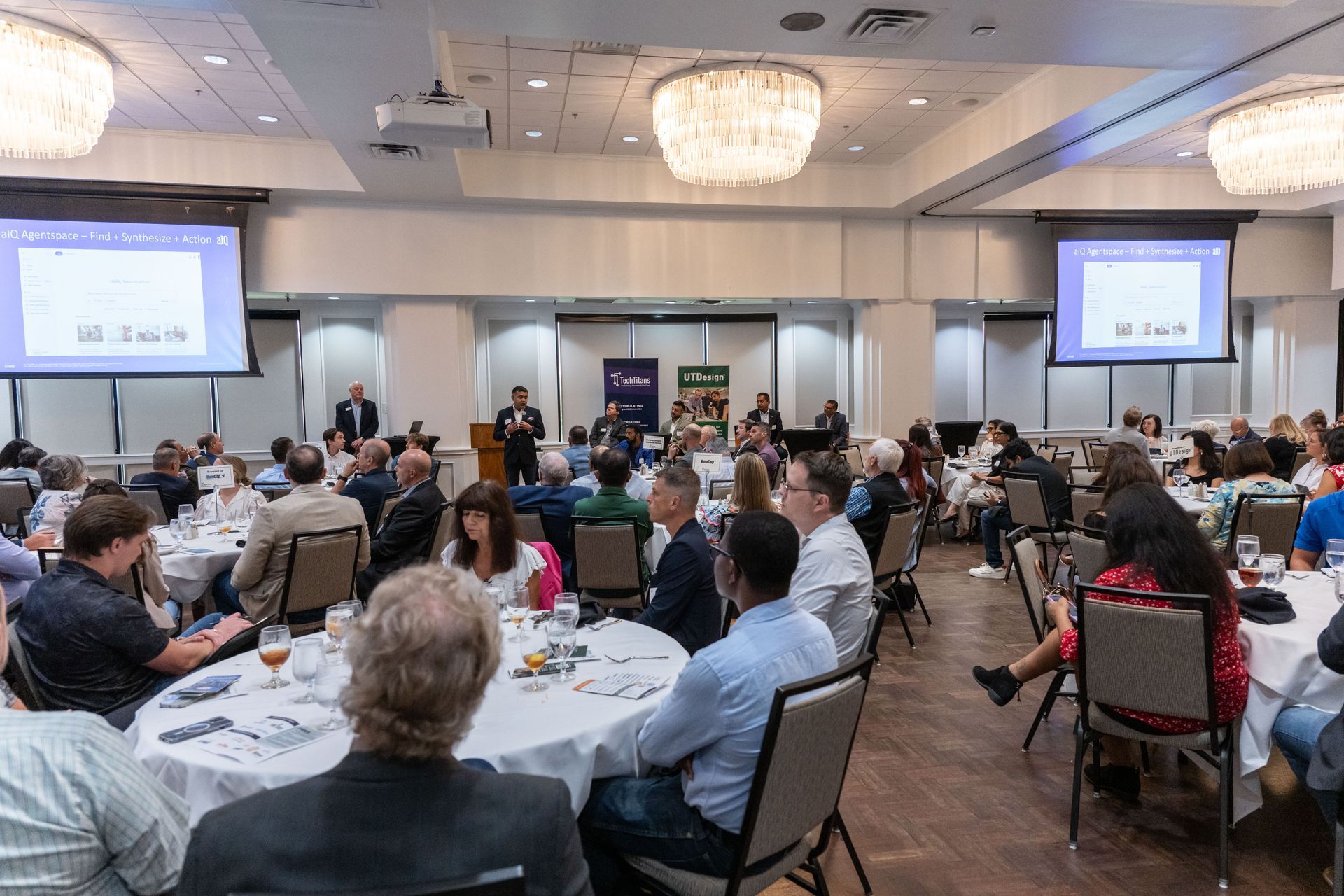 Attendees seated at round tables in a conference room listening to a presentation with projector screens on either side.