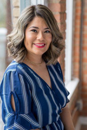 A smiling person with wavy brown hair wearing a blue and white striped shirt, standing near a brick wall and window.