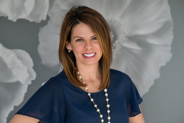 A person with shoulder-length brown hair wears a navy blue top and a pearl necklace, smiling against a floral backdrop.