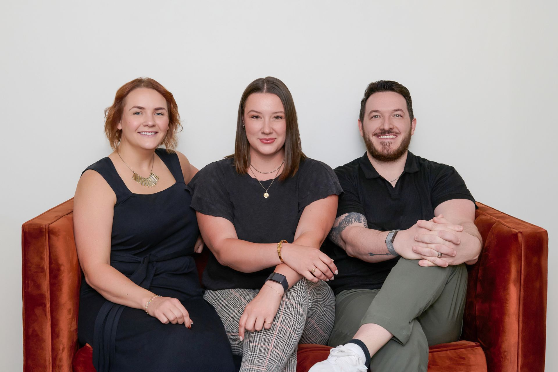 Three people sit smiling on an orange sofa against a white wall.