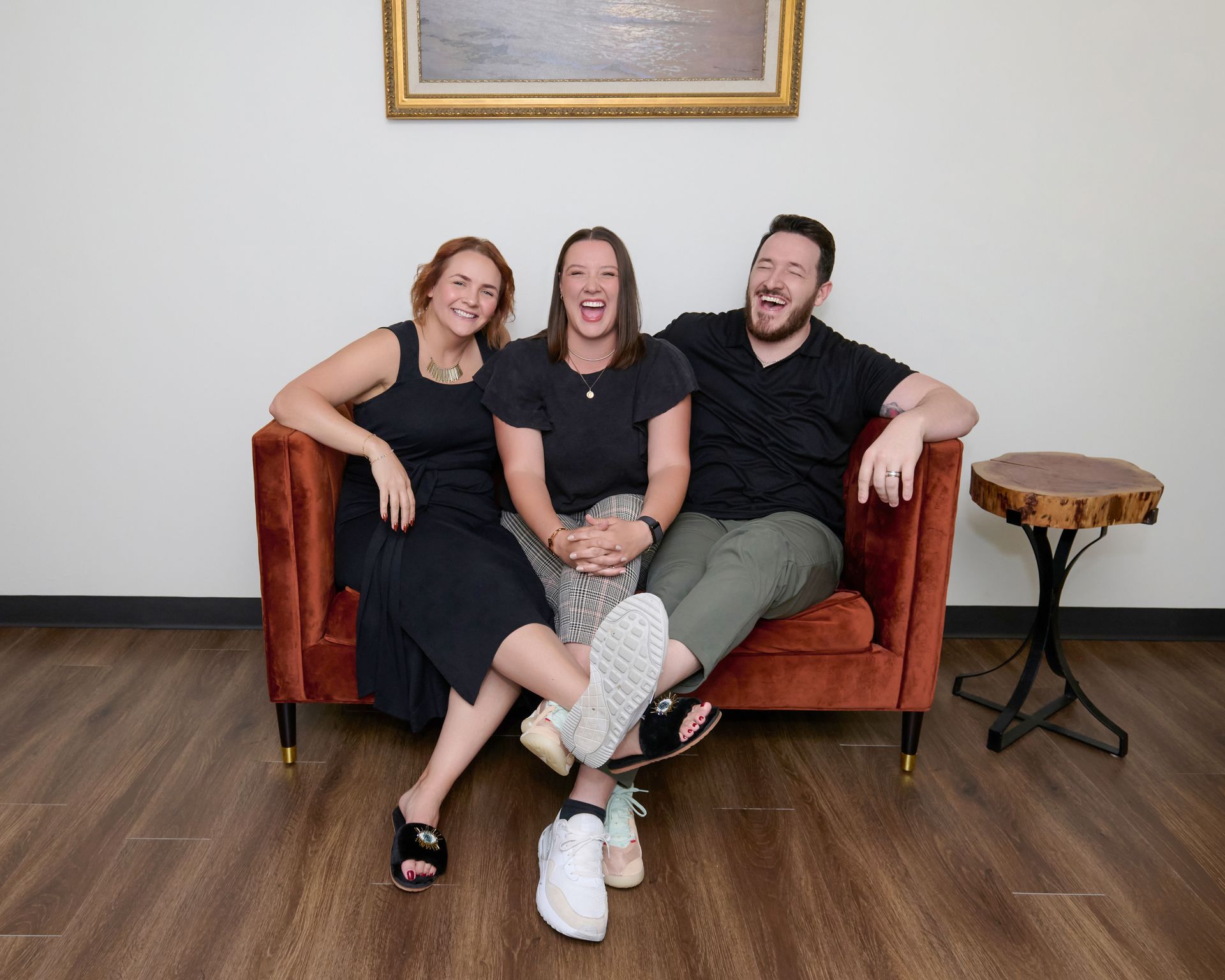 Three people laughing while sitting on a burnt orange velvet couch against a white wall with a framed painting.