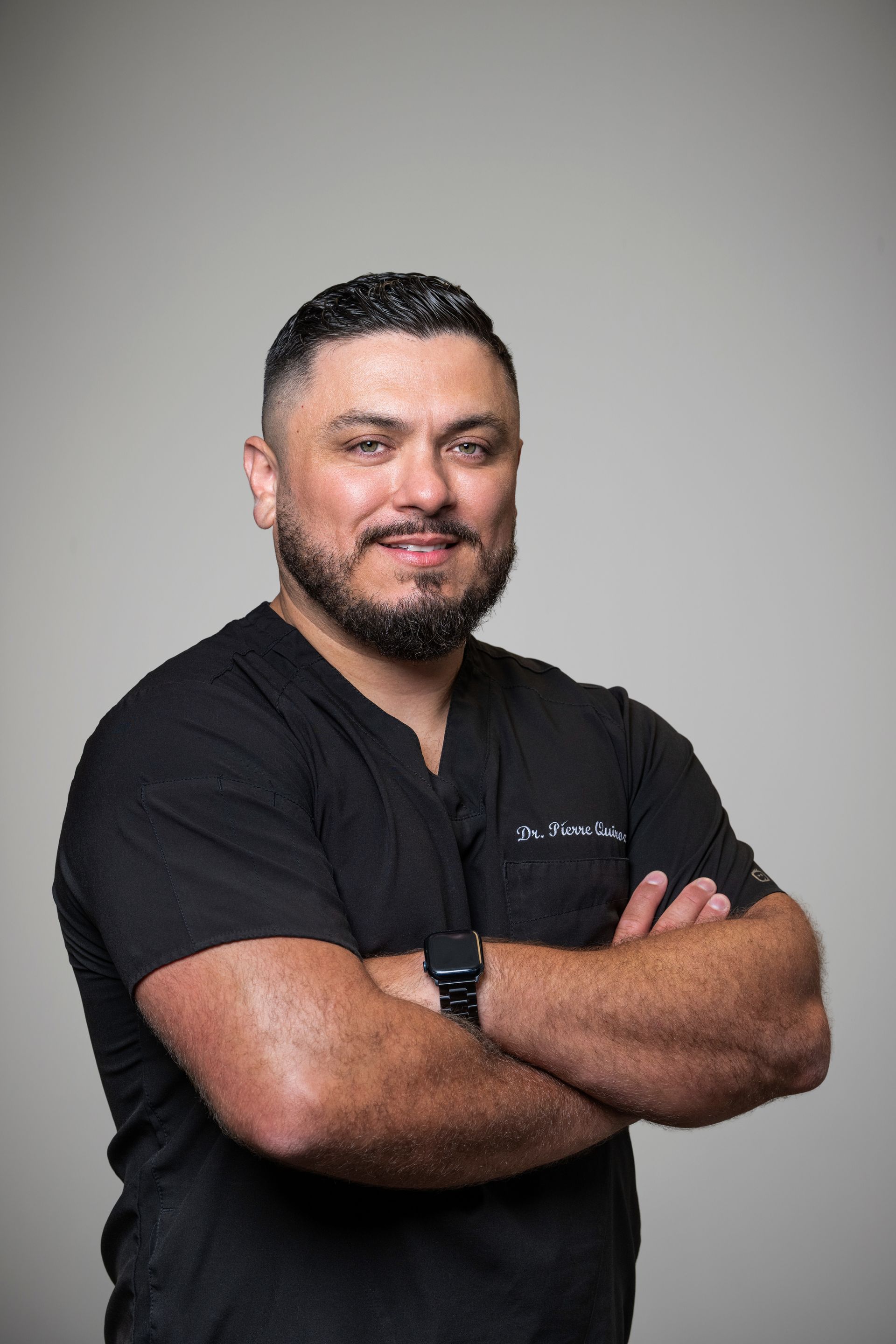 A person with a beard, wearing a black scrub top, stands against a plain background with arms crossed, smiling slightly.