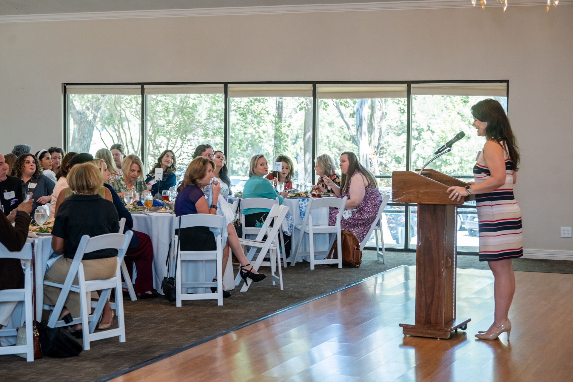 A presenter speaks from a wooden podium to an audience seated at tables in a brightly lit, spacious function hall.