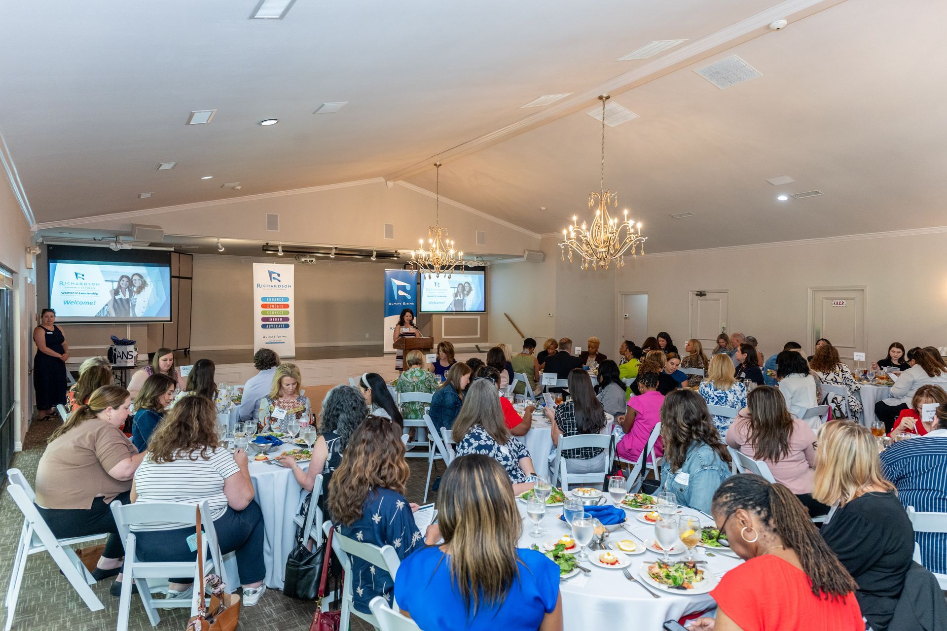 A presentation is held in a ballroom with guests seated at round tables facing a stage and projector screens.
