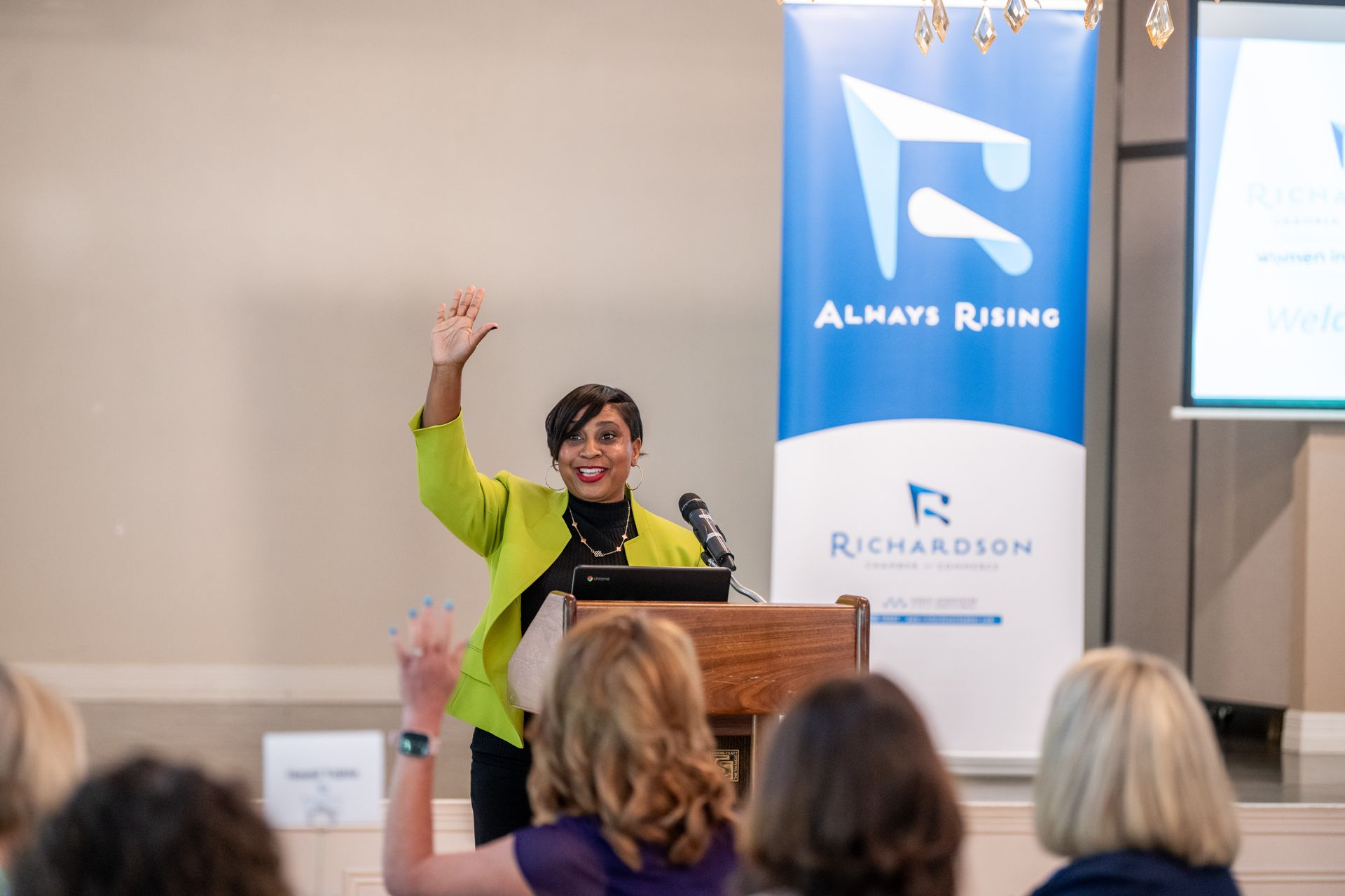 A woman in a bright lime jacket speaks at a podium with a blue 