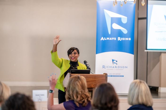 A speaker in a lime green blazer stands at a podium, waving to an audience, with an Always Rising banner in the background.
