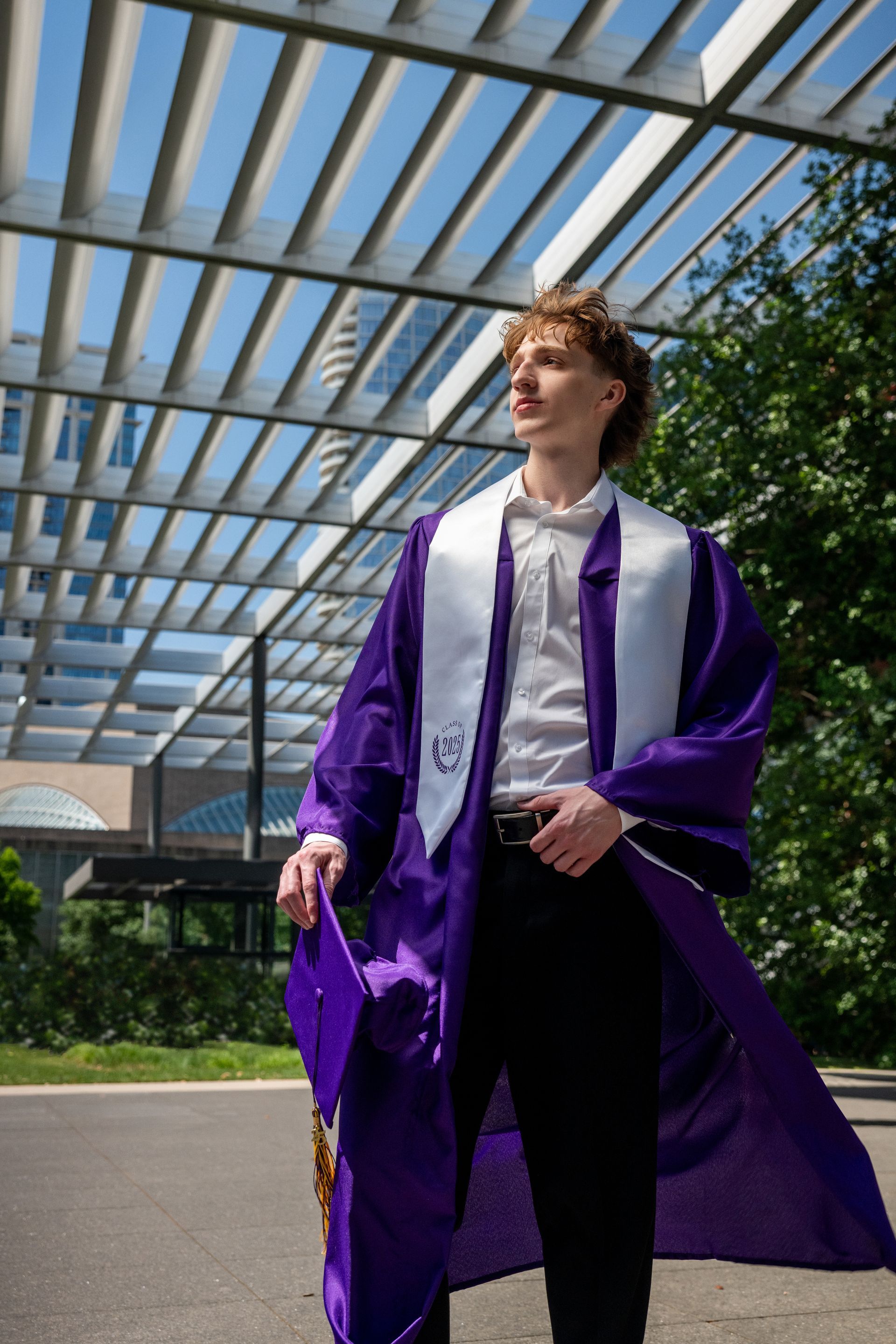 A person in a purple graduation gown and white stole stands under a slatted architectural canopy outdoors.