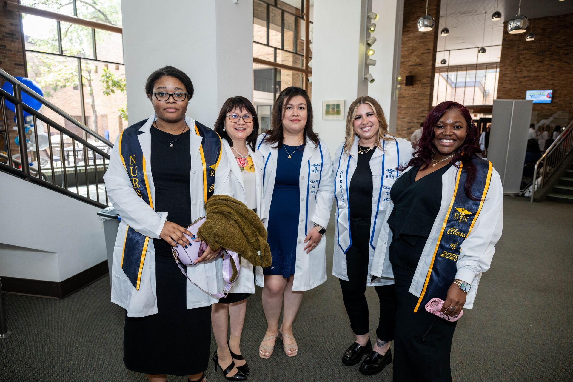 Five people in professional attire and white clinical coats pose for a photo indoors, some wearing graduation stoles.