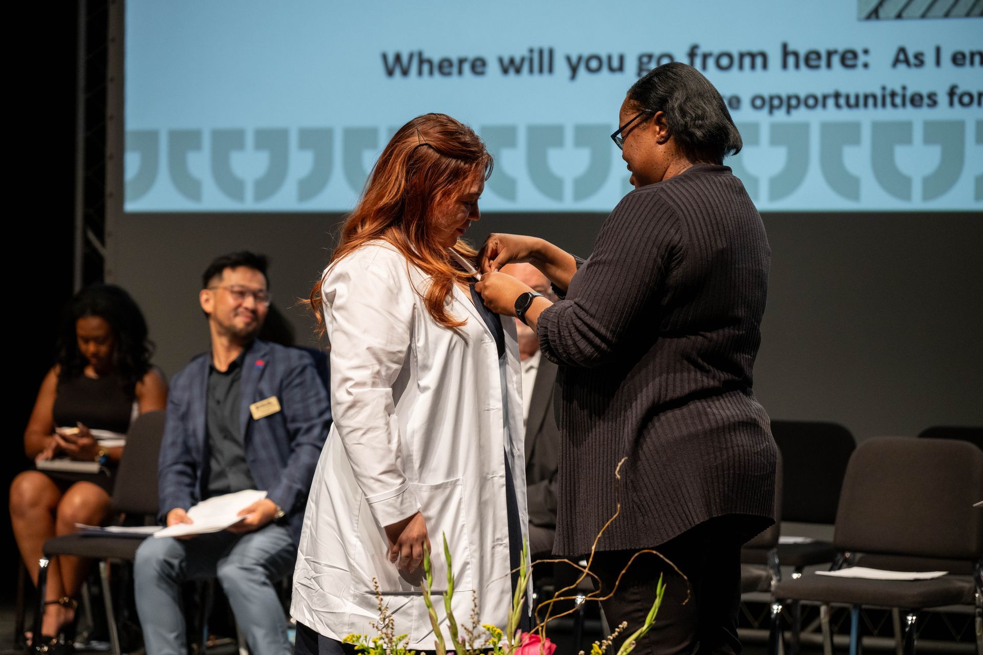 A person in a white coat has a pin attached to their lapel by another person on a stage during an event.