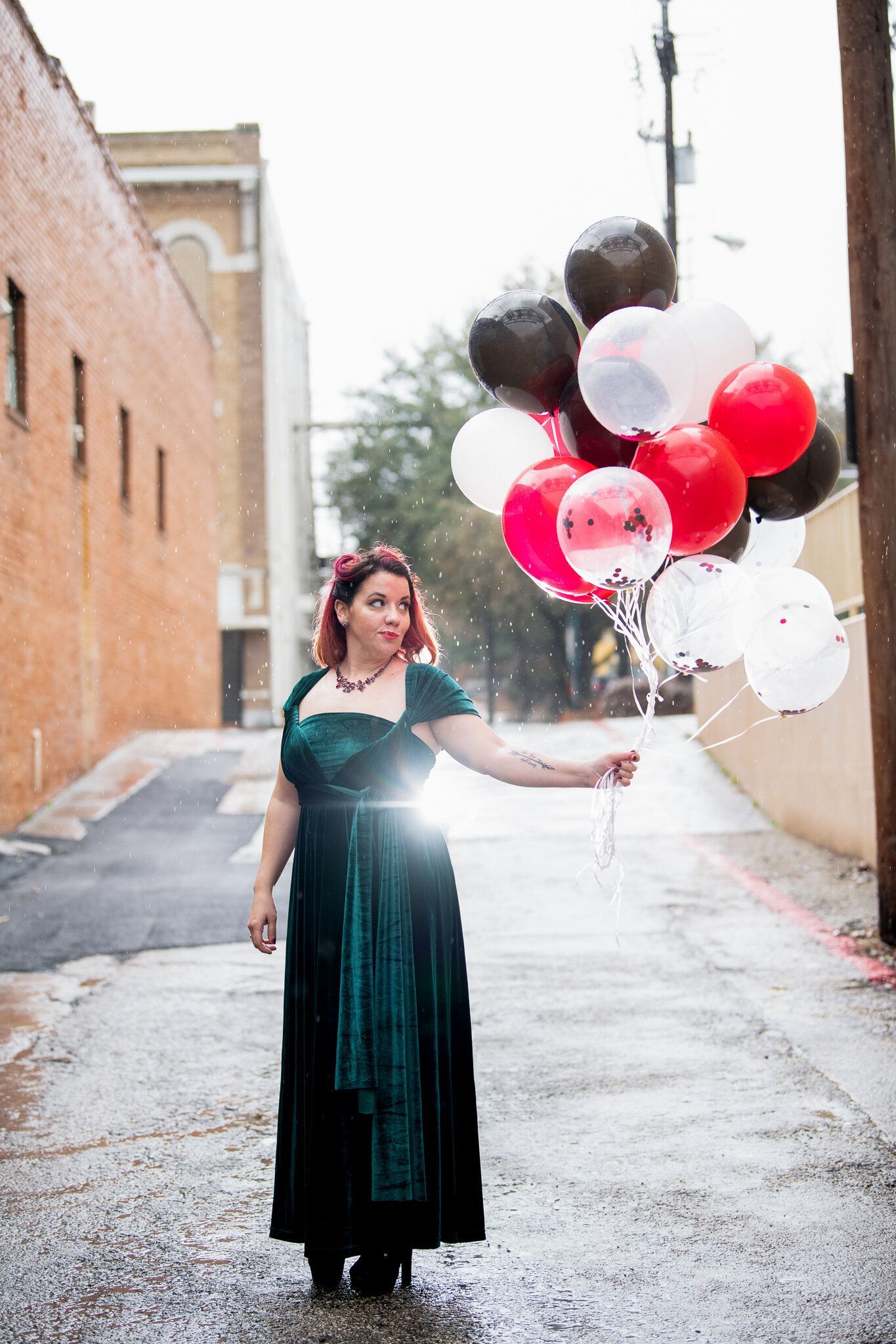 A person in a dark green velvet dress stands in an alleyway holding a cluster of red, white, and black balloons.