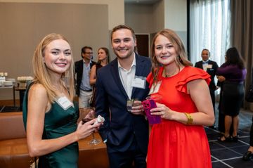 Three people smile while socializing at an indoor networking event. They hold drinks and wear business attire.