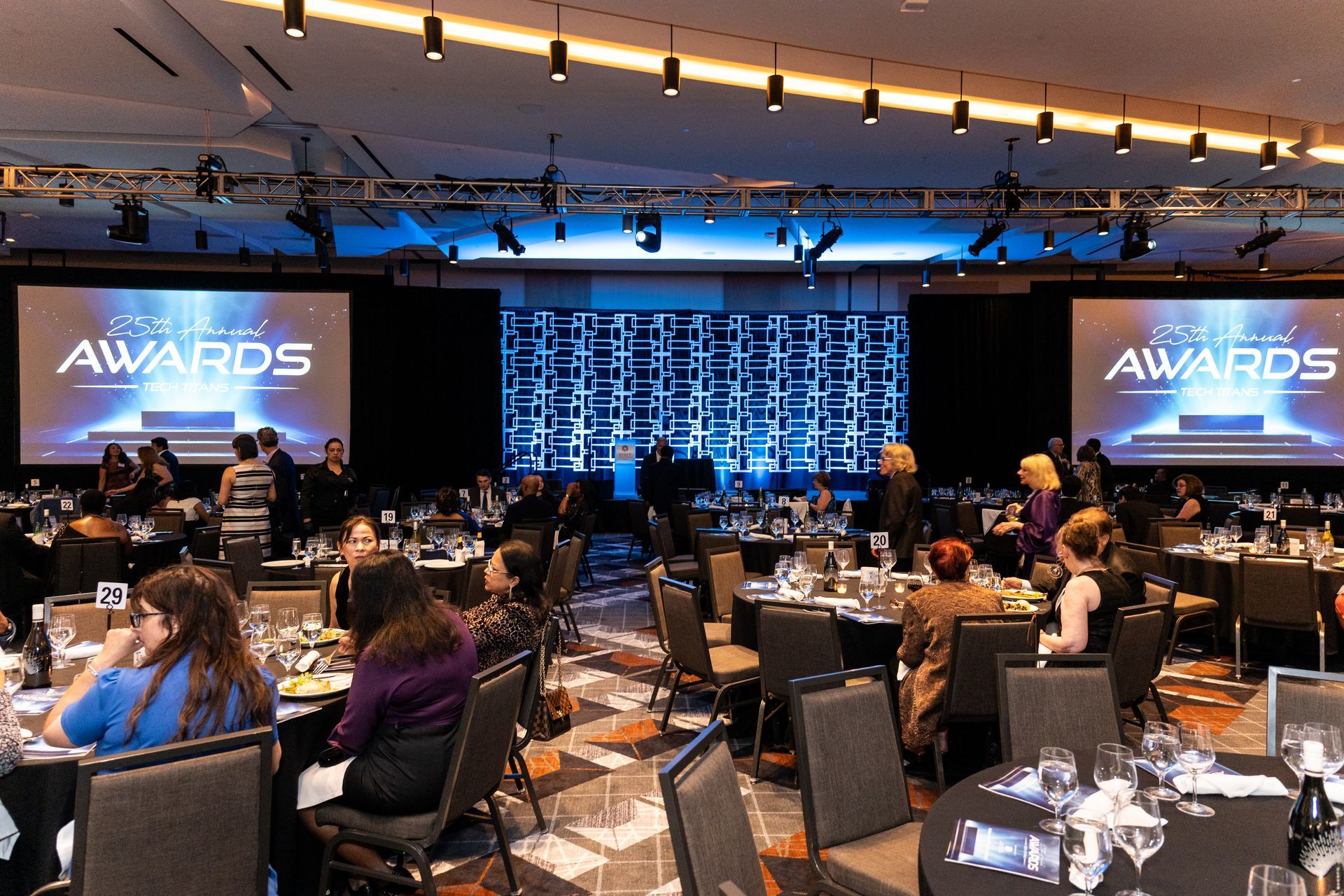 A banquet hall for an awards event with round tables, attendees, and large screens displaying 