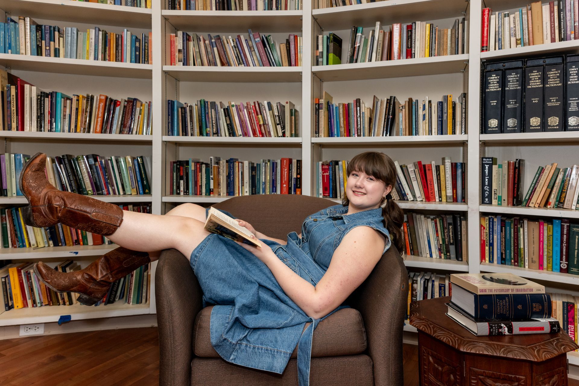 A person in a denim dress and brown cowboy boots reclines in a chair, reading a book in front of a wall of bookshelves.