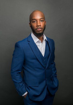 A man wearing a blue three-piece suit and white shirt, standing with hands in his pockets against a gray background.