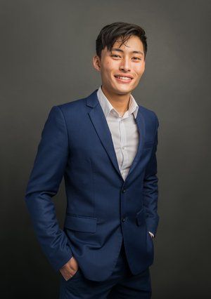 A person in a blue suit and white dress shirt smiling, hands in pockets, standing against a dark grey background.