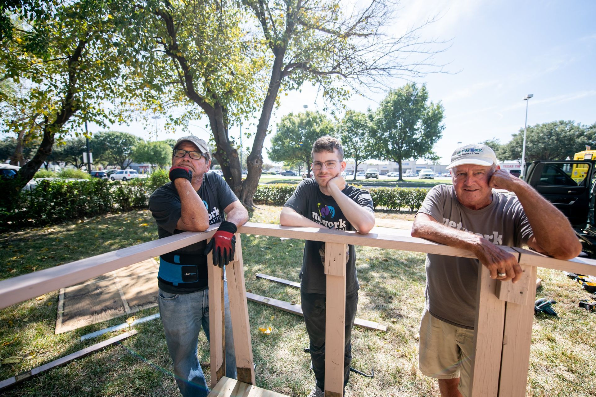 Three people lean on a wooden frame outdoors, looking at the camera in a sunny, grassy area with trees and a parked truck.
