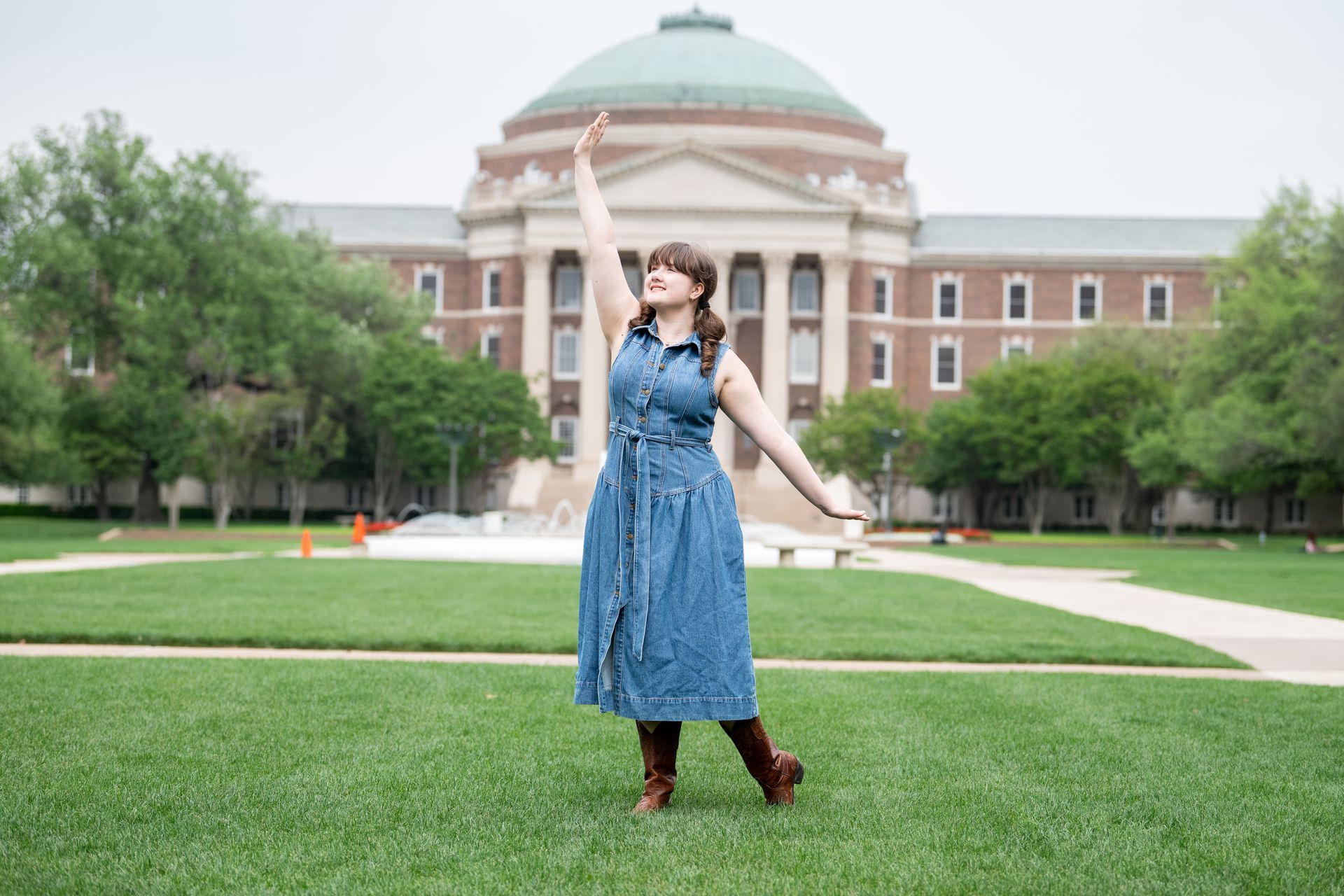 A person wearing a denim dress and boots poses with one arm raised in front of a domed university building on a lawn.