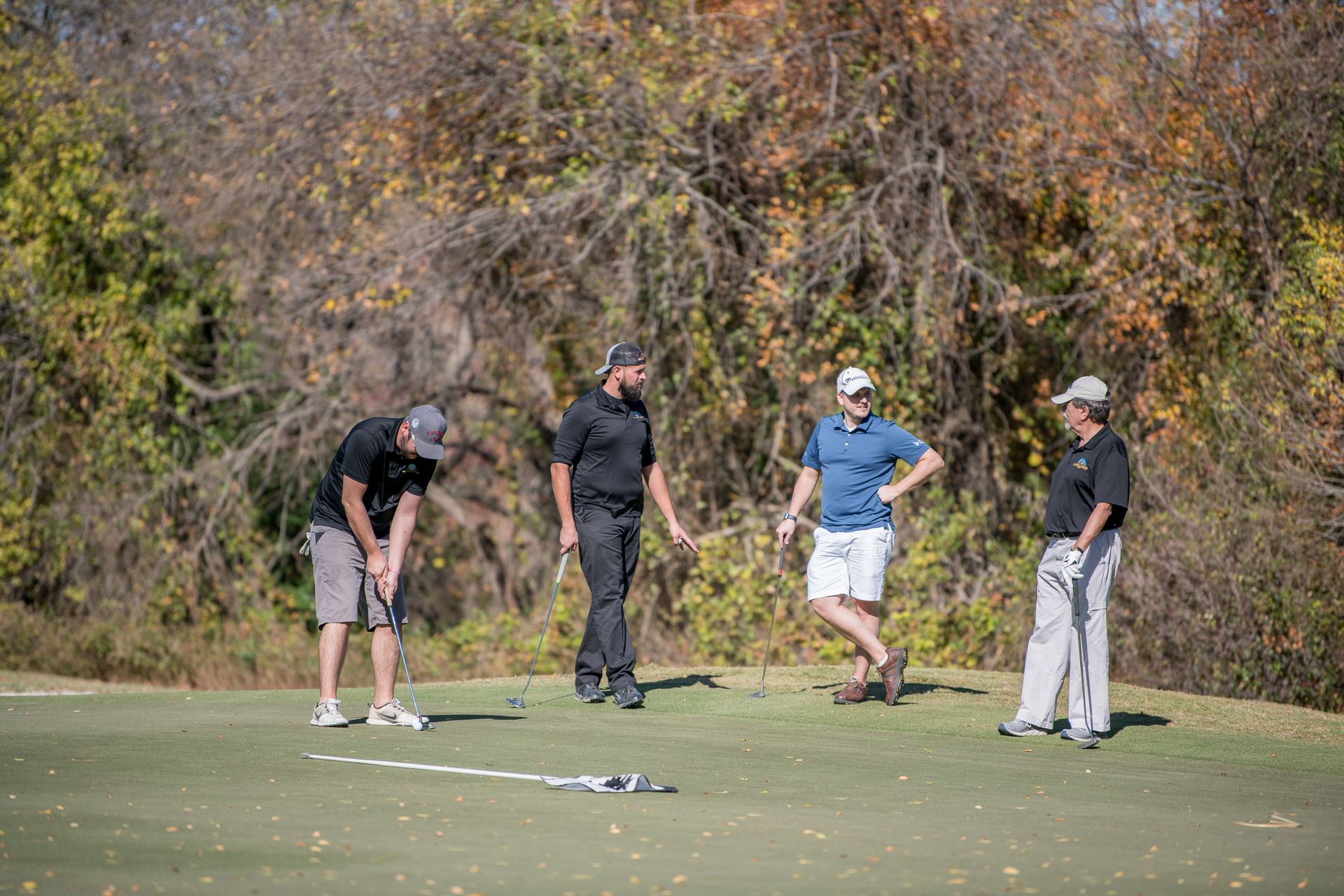 Four people on a putting green, one preparing to putt while the others watch in a wooded outdoor setting.