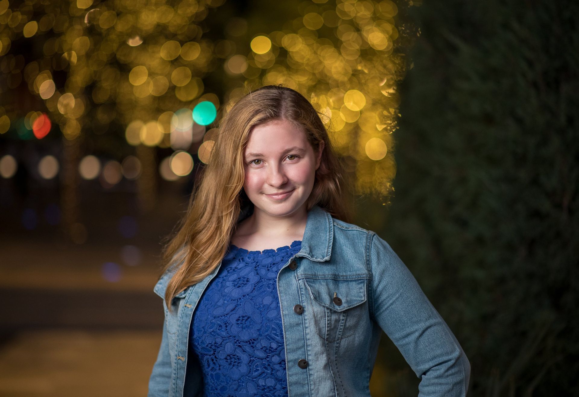 Smiling individual wearing a blue lace top and denim jacket, posed against a blurry, festive city light background.