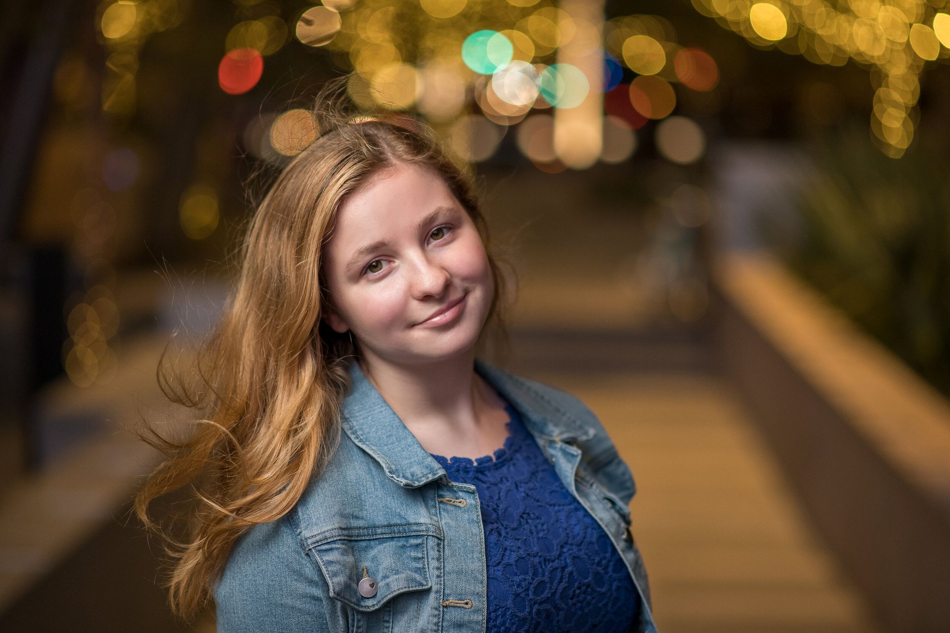 A smiling person with long light brown hair wears a blue top and denim jacket against a blurred, glowing city light backdrop.