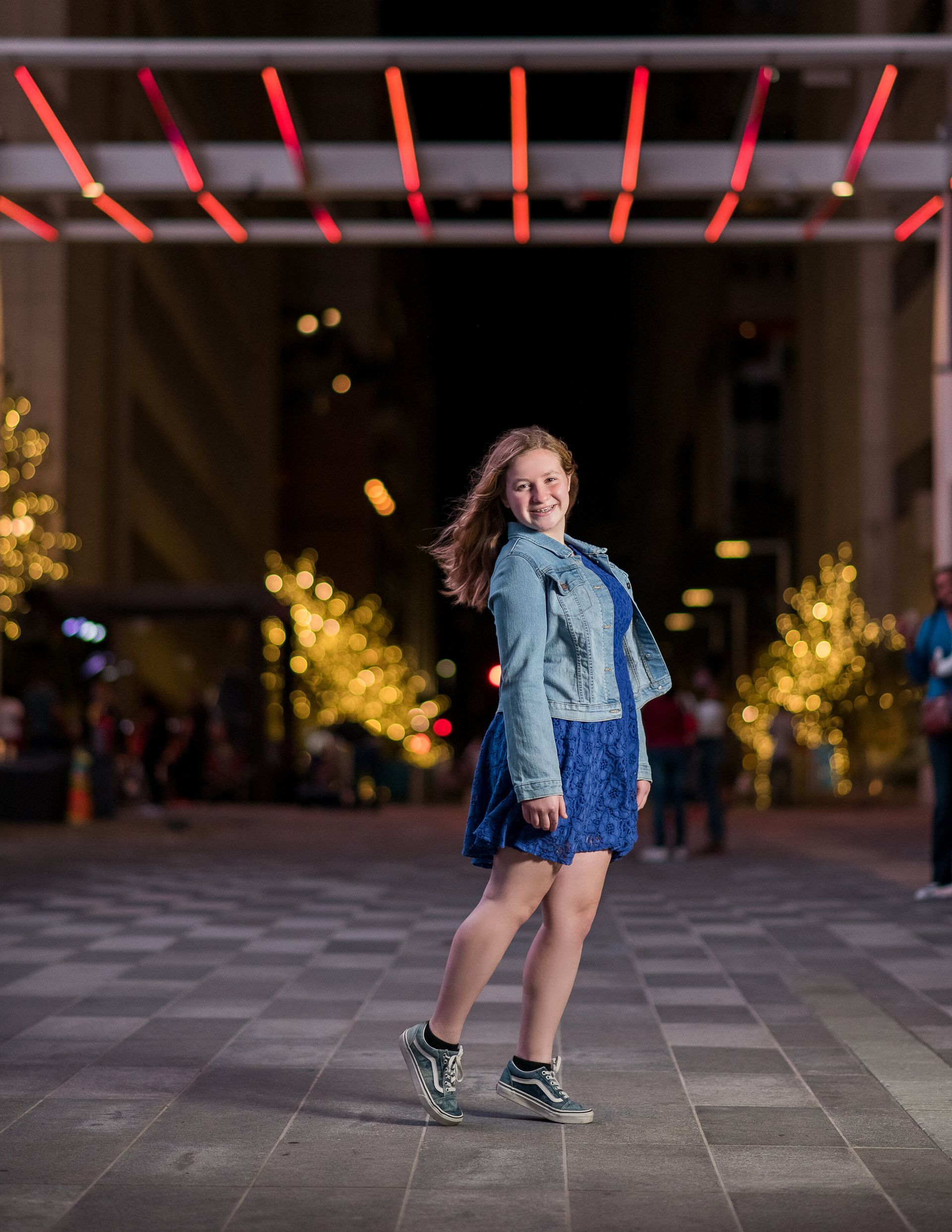A person in a denim jacket and blue patterned dress poses smiling on a paved urban plaza illuminated by festive lights.