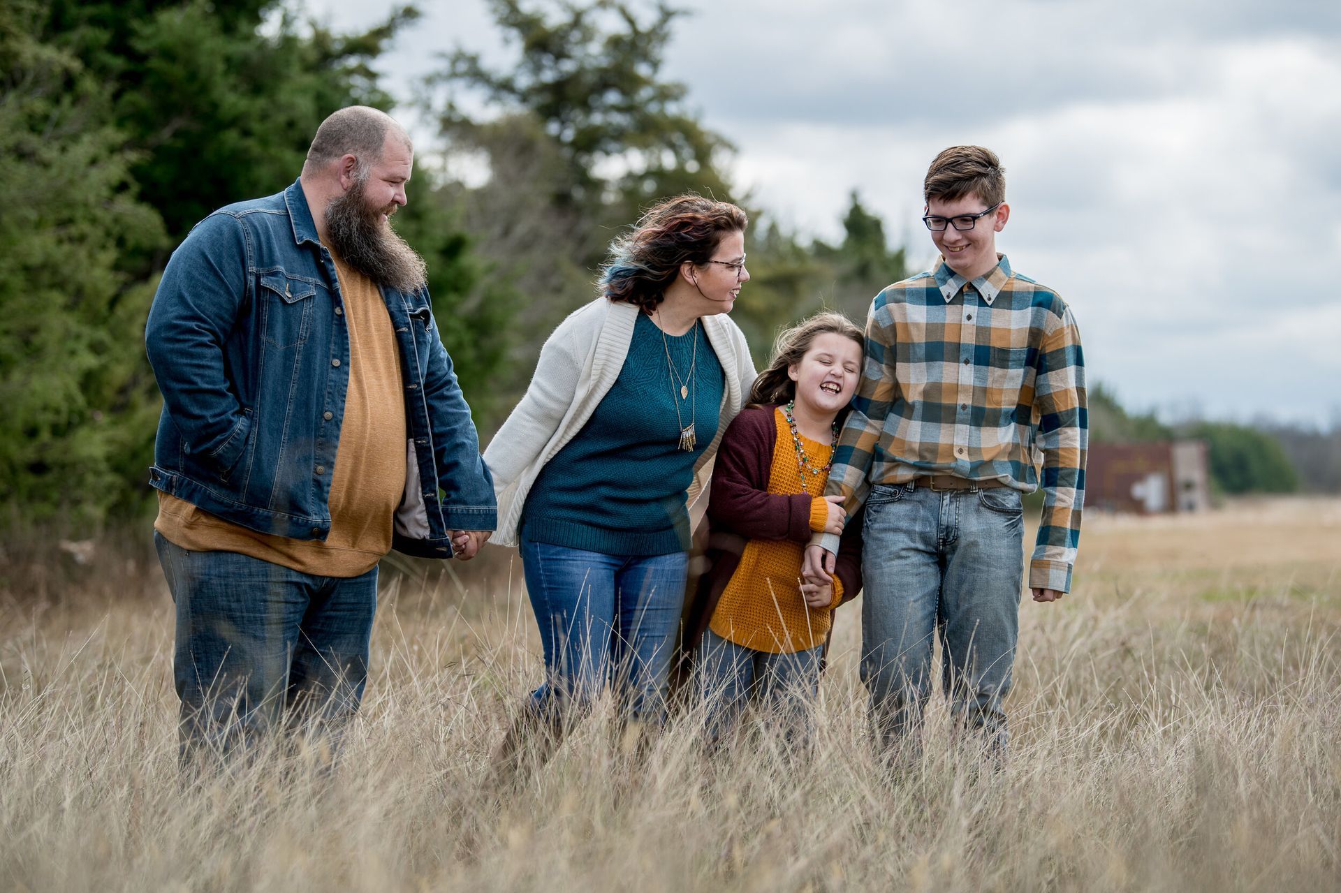 A family of four walking through a dry, grassy field toward the right, smiling and laughing under an overcast sky.