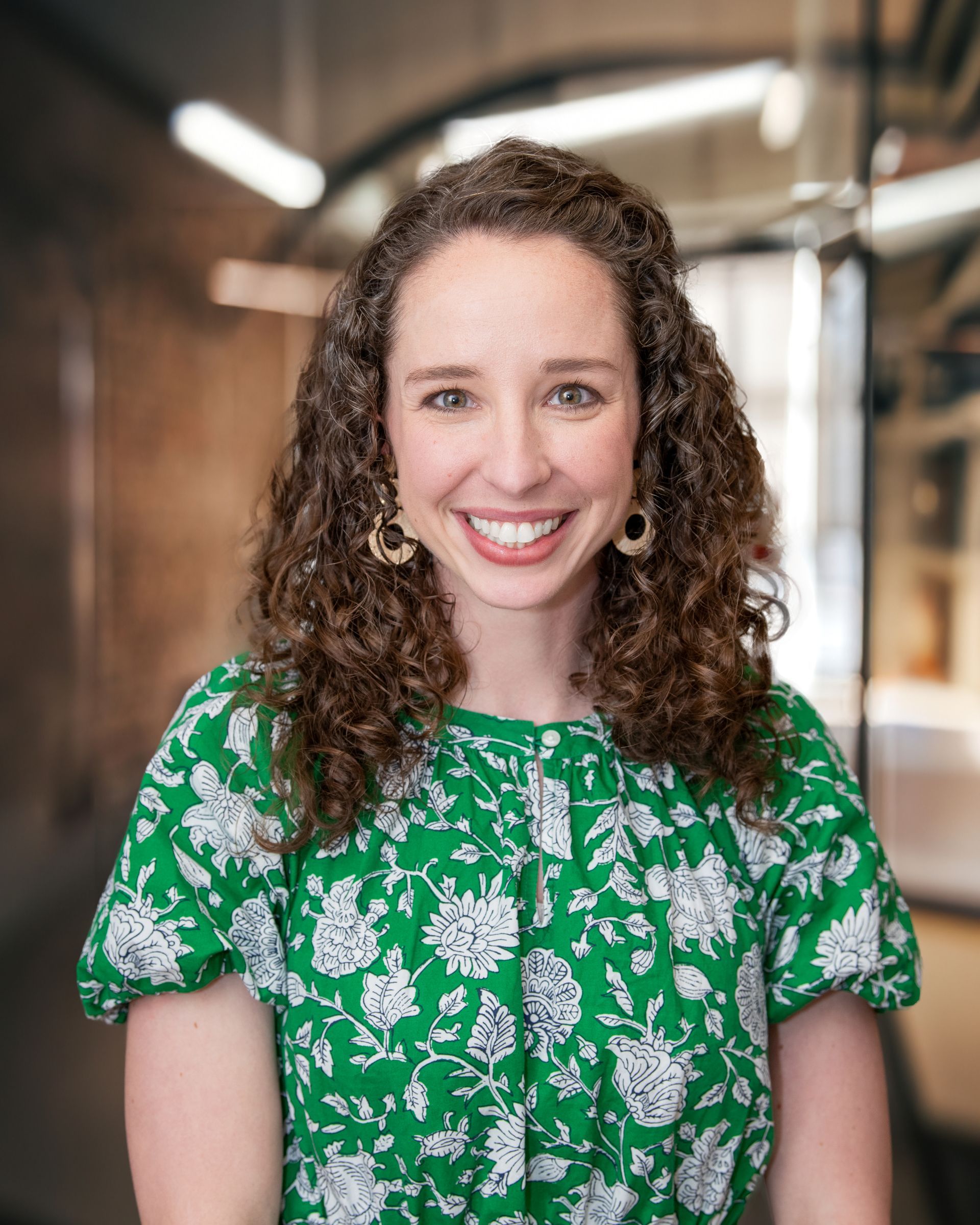 A smiling person with curly brown hair wearing a green and white floral patterned top in an office setting.