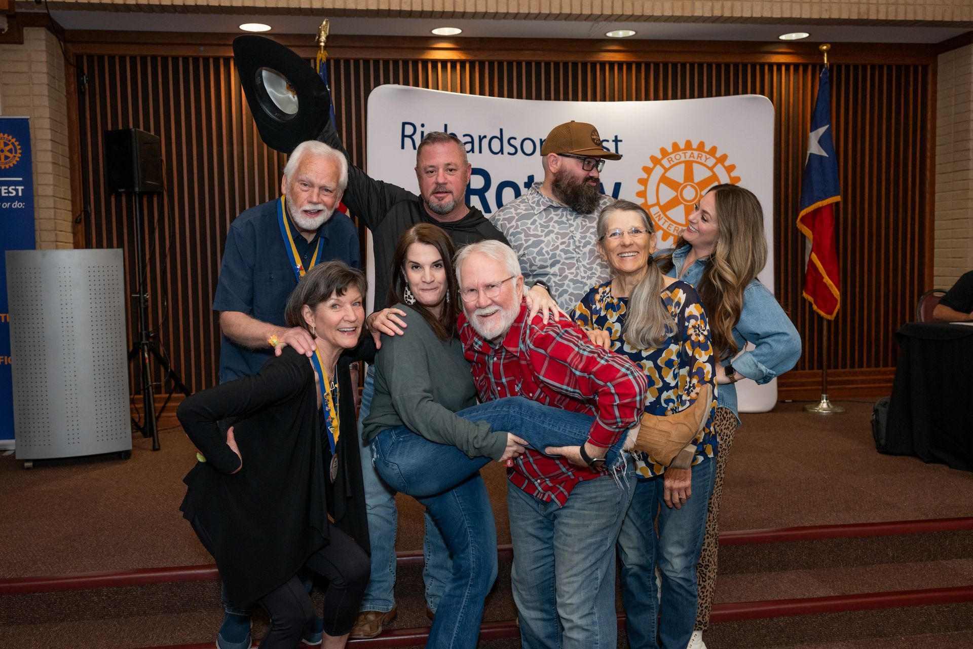 A group of eight people pose for a playful photo in front of a Rotary Club banner and a Texas state flag.