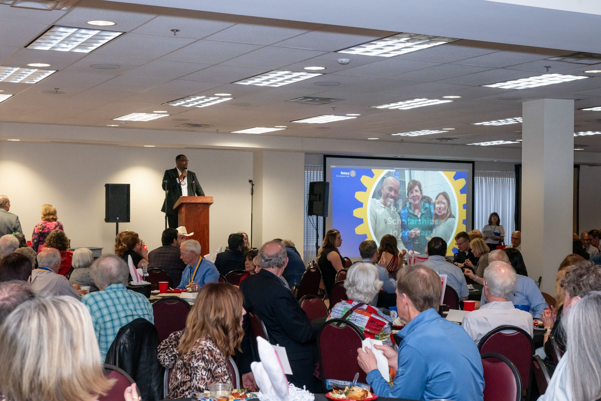 A speaker stands at a podium in a conference room addressing an audience facing a large screen displaying a group photo.