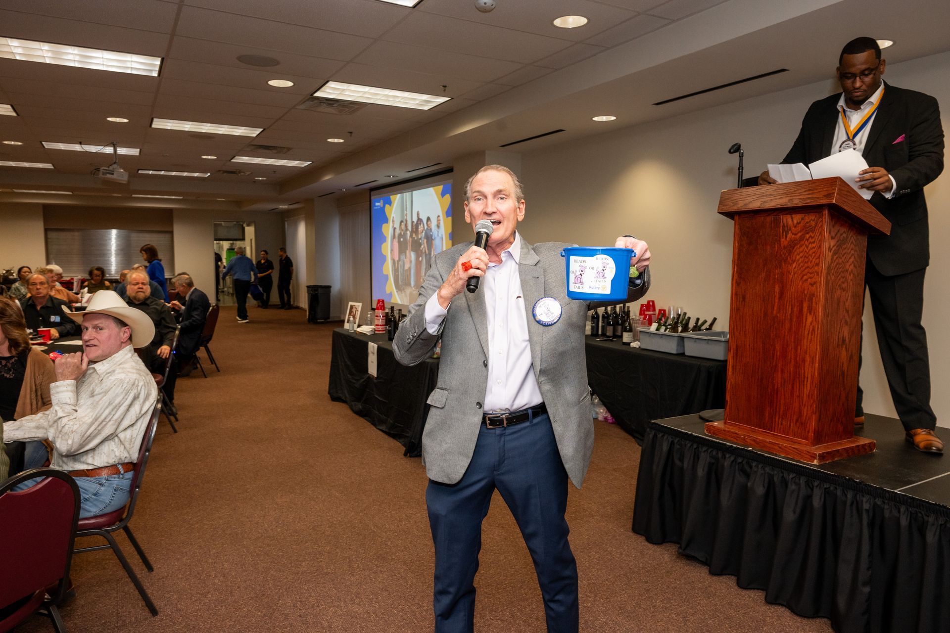 A speaker holds a small blue bucket while addressing a seated audience from an indoor event space with a wooden podium.