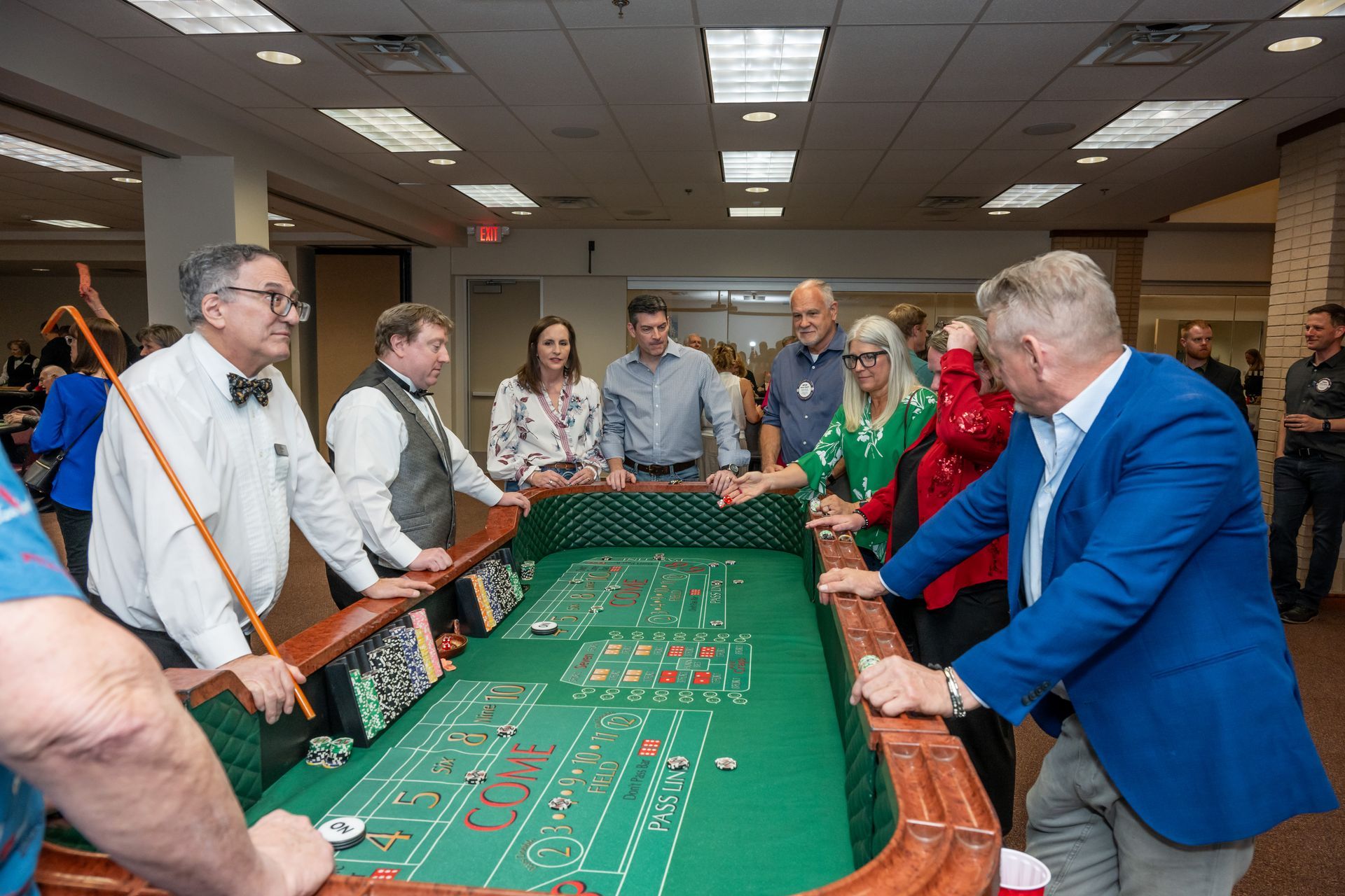 A group of people standing around a green craps table, interacting in an indoor setting with overhead lighting.