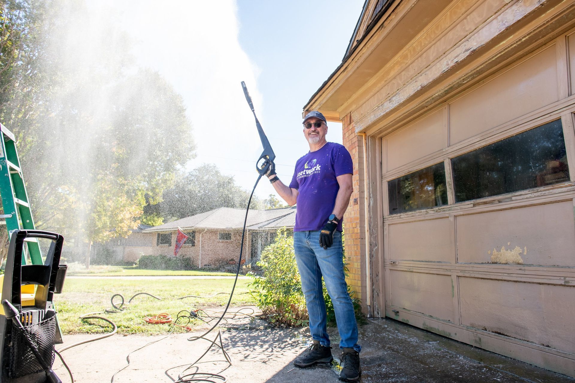 A person in a purple shirt uses a pressure washer to clean the side of a garage on a sunny day.