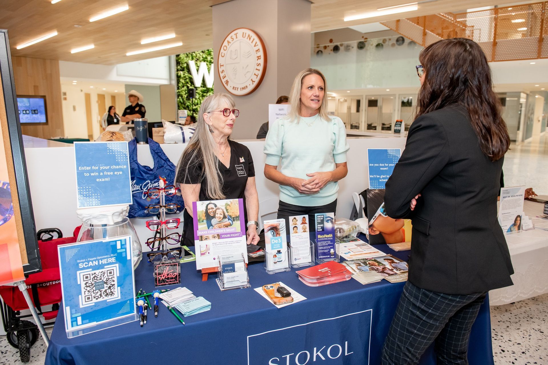 Three people stand at a table draped in a blue cloth, engaging in conversation at an indoor university event.