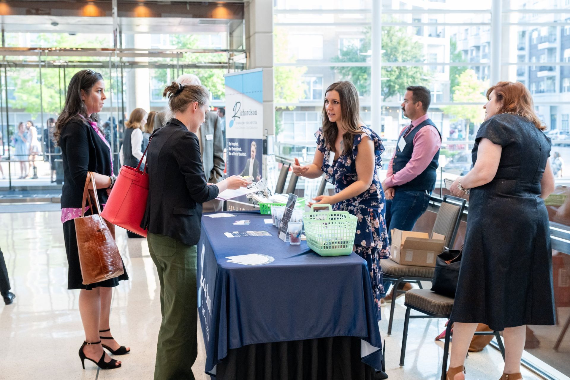 People in business casual attire interact at an information table during a professional conference in a bright lobby.