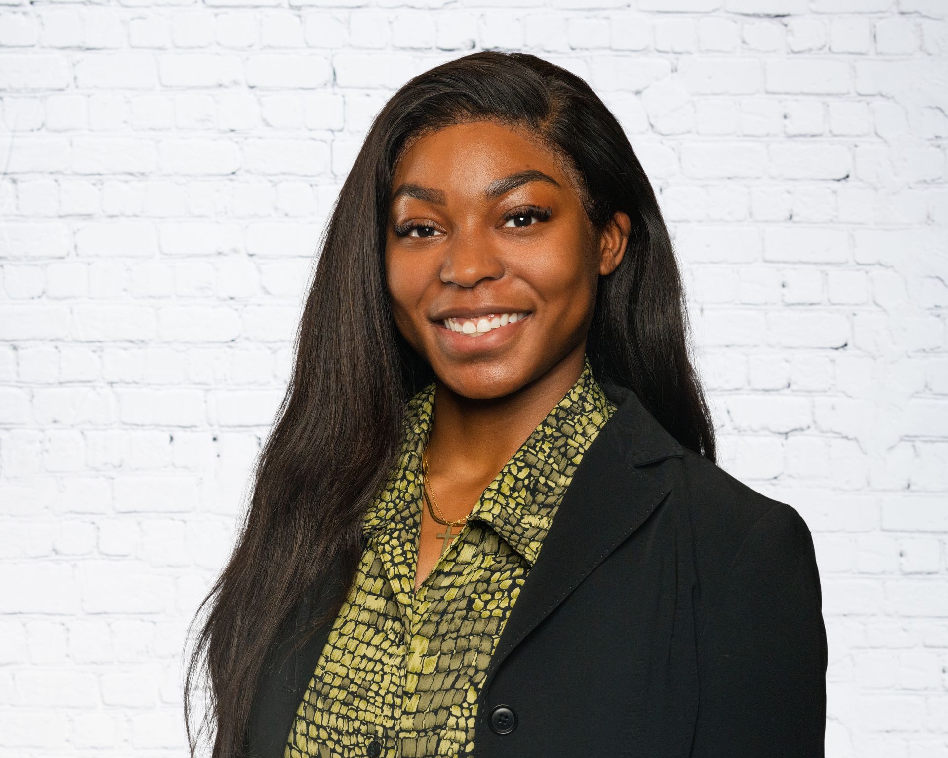 A smiling person with long dark hair wears a black blazer over a patterned green shirt against a white brick wall.