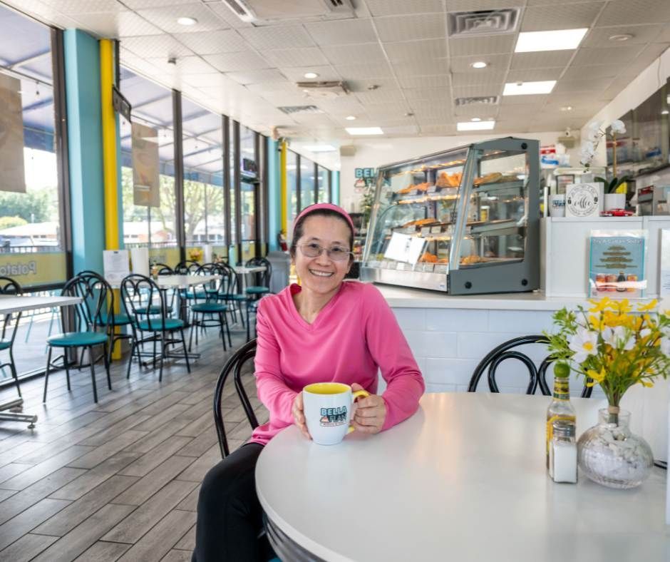 A smiling person wearing a pink sweater sits at a cafe table holding a mug, with a pastry display case in the background.