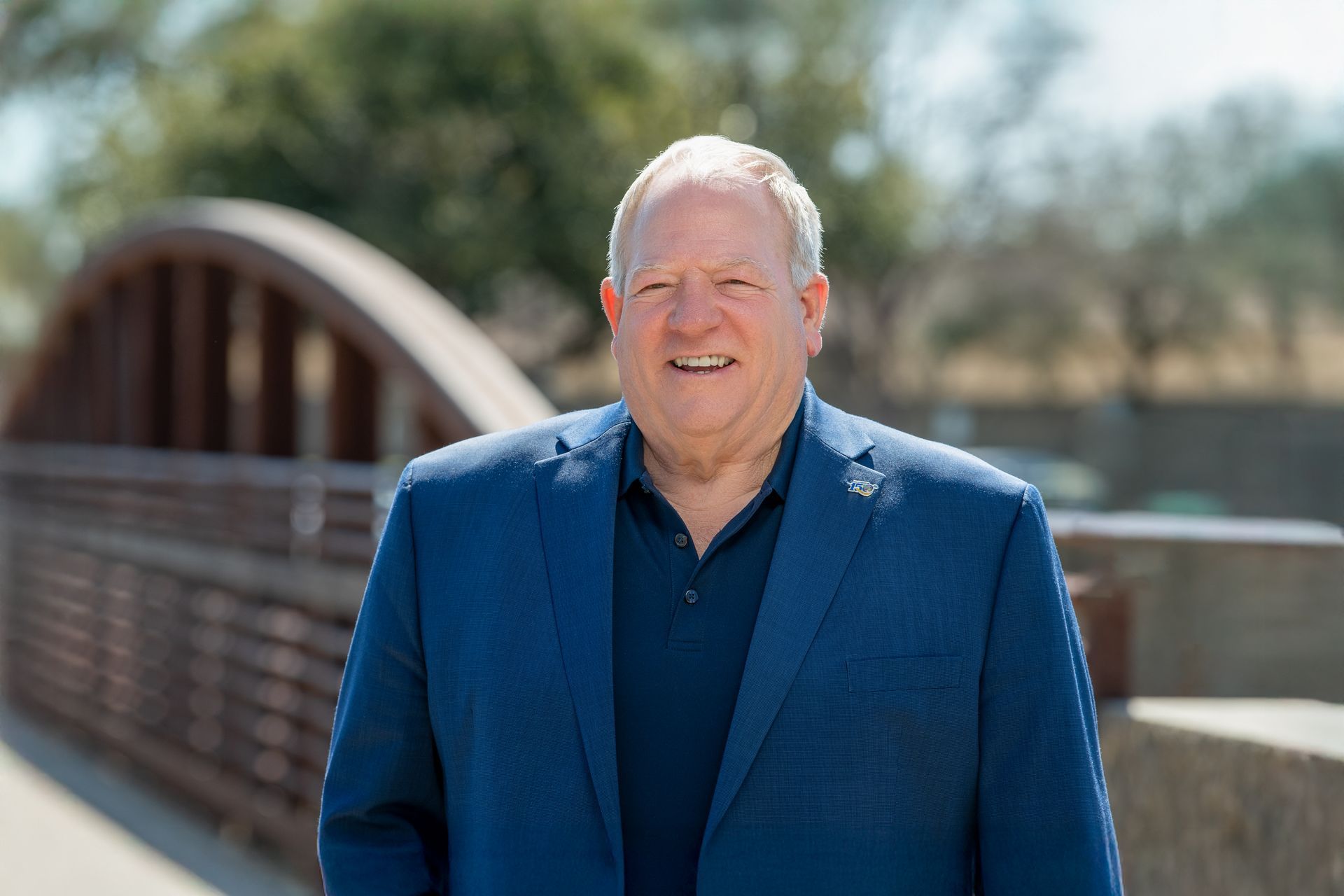 A smiling person in a blue blazer and dark shirt stands outdoors on a pedestrian bridge with a wooden railing.