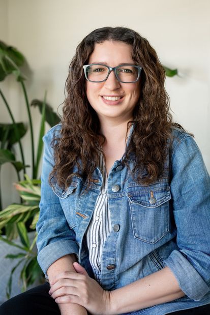 A person with long, curly brown hair and glasses wears a blue denim jacket, smiling in front of green leafy plants.