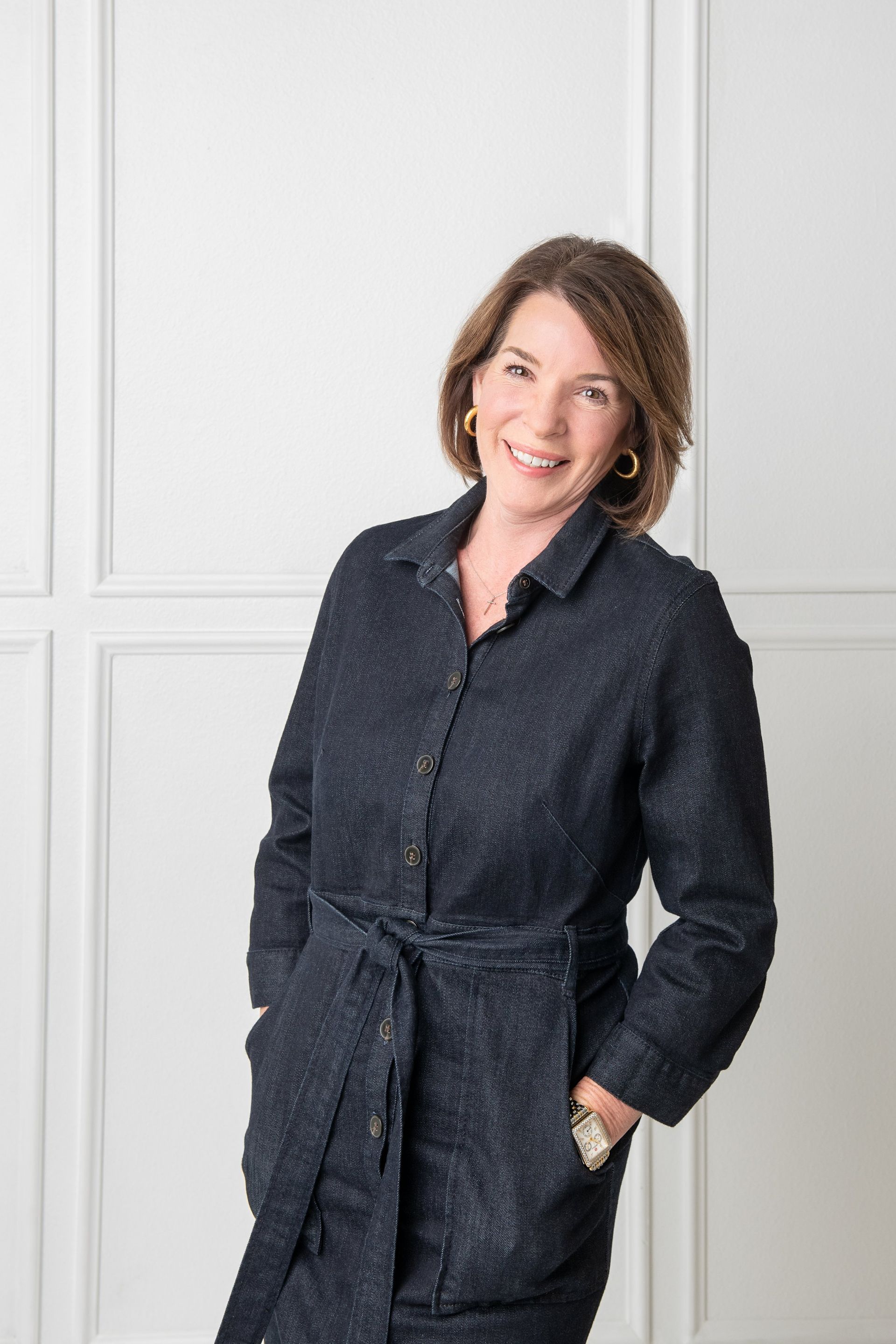 A professional portrait of a person smiling, wearing a dark-textured, belted shirt dress against a white paneled wall.