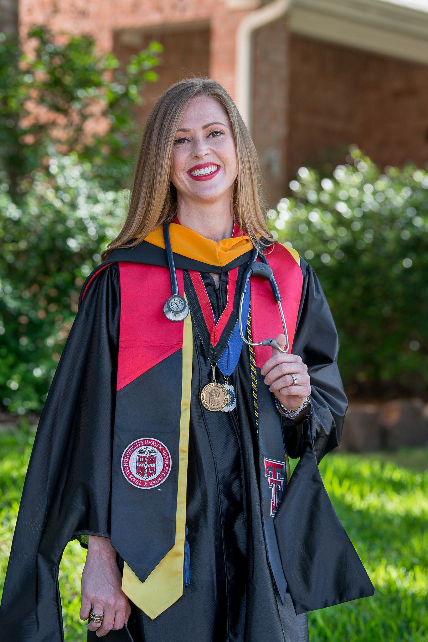 A smiling graduate in academic regalia, including a black robe and a red and gold stole, stands outdoors in a green area.
