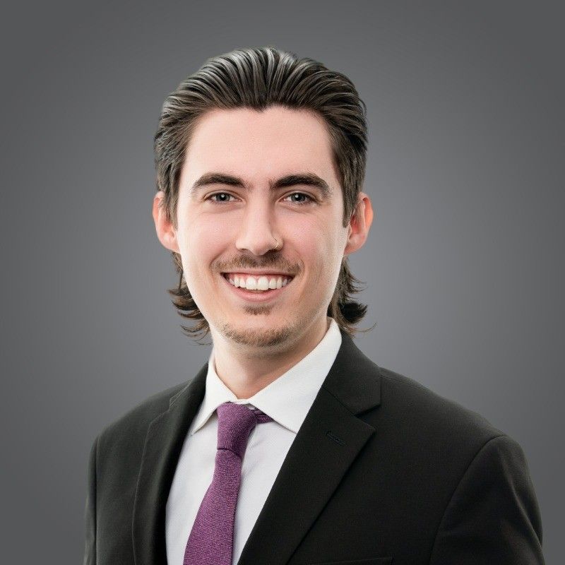 Headshot of a person with dark hair, smiling while wearing a black suit, white shirt, and purple tie against a grey background.