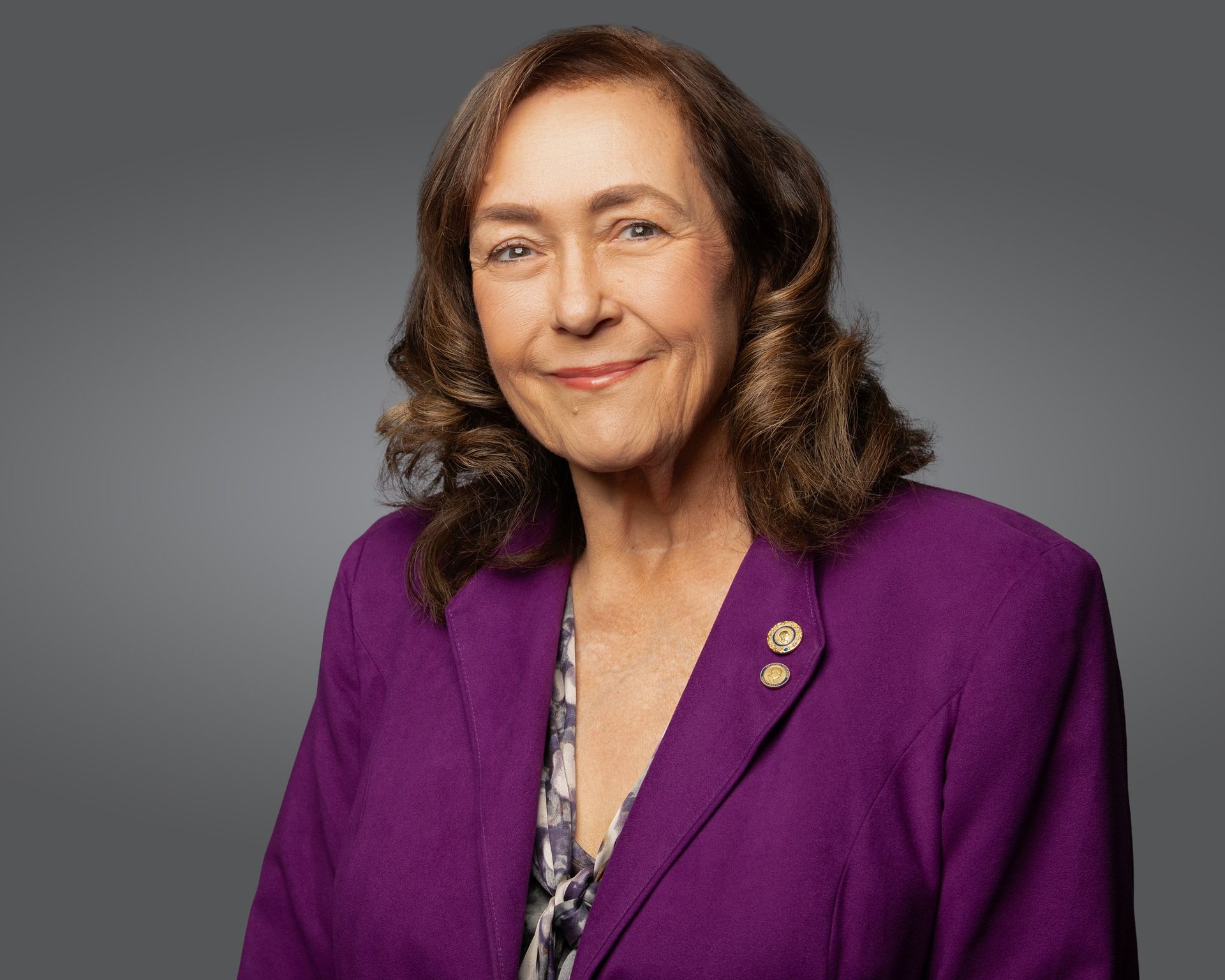 A person with wavy brown hair wearing a purple blazer over a patterned top, smiling against a gray background.