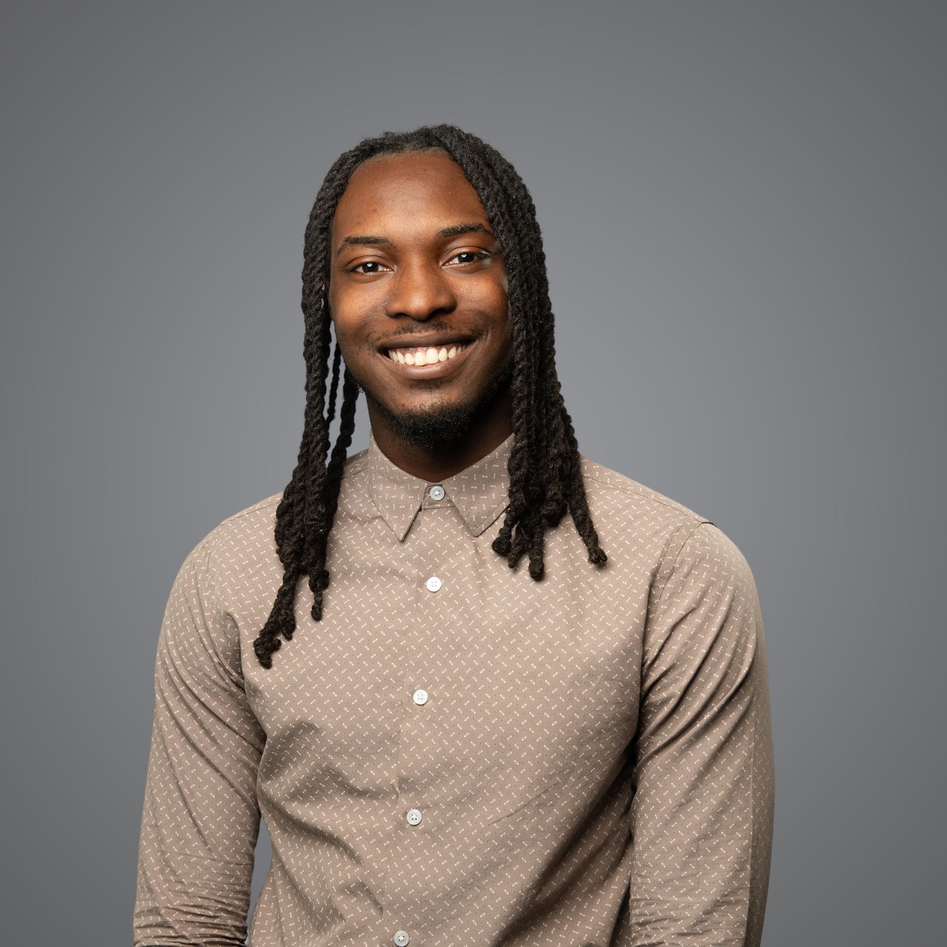 A person with long braided hair wearing a light brown, patterned button-down shirt, smiling against a gray background.