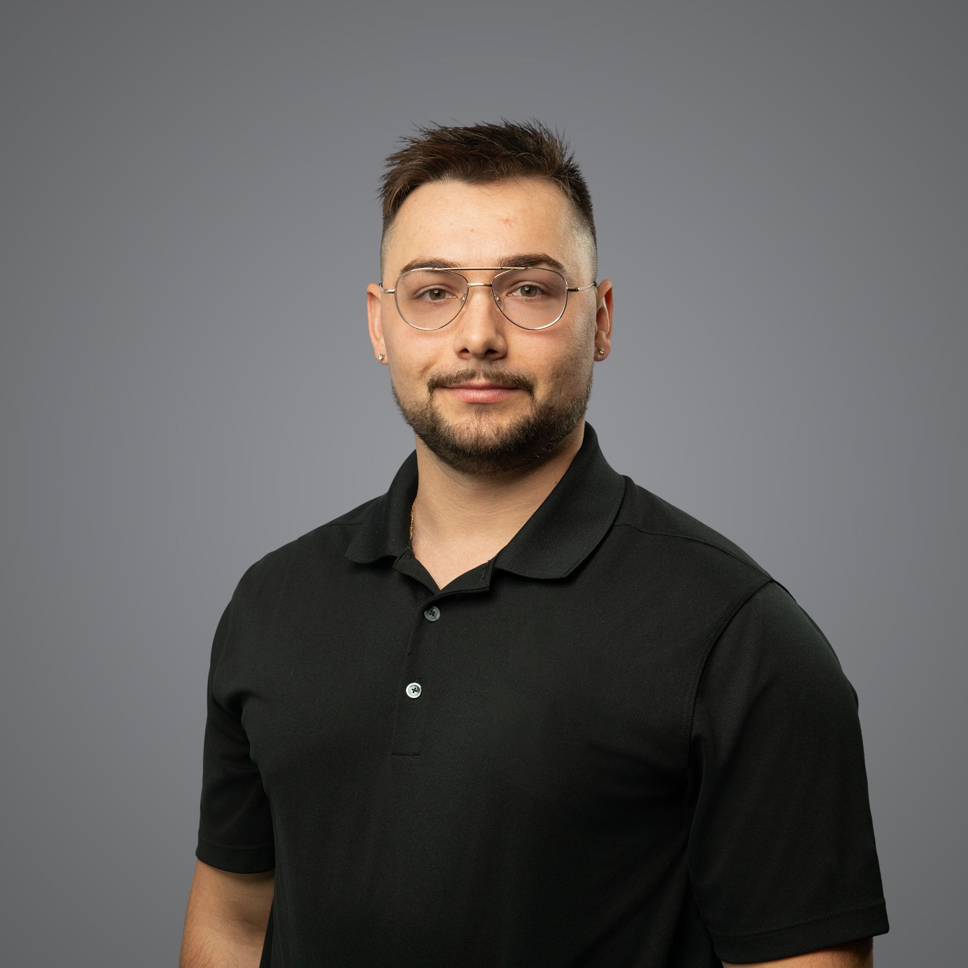 A person wearing glasses and a black polo shirt, posing for a portrait against a solid gray background.